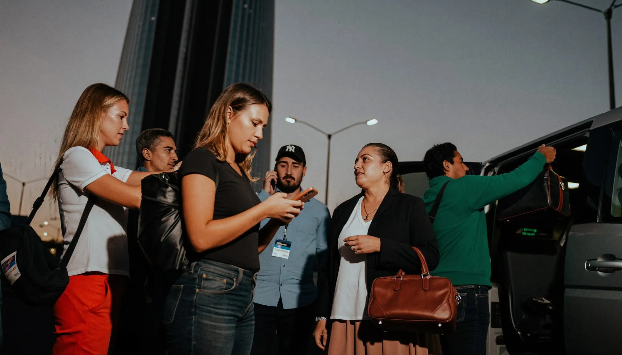 Colleagues with overnight bags waiting beside a vehicle at dusk outside a Dubai office tower, one checking a phone