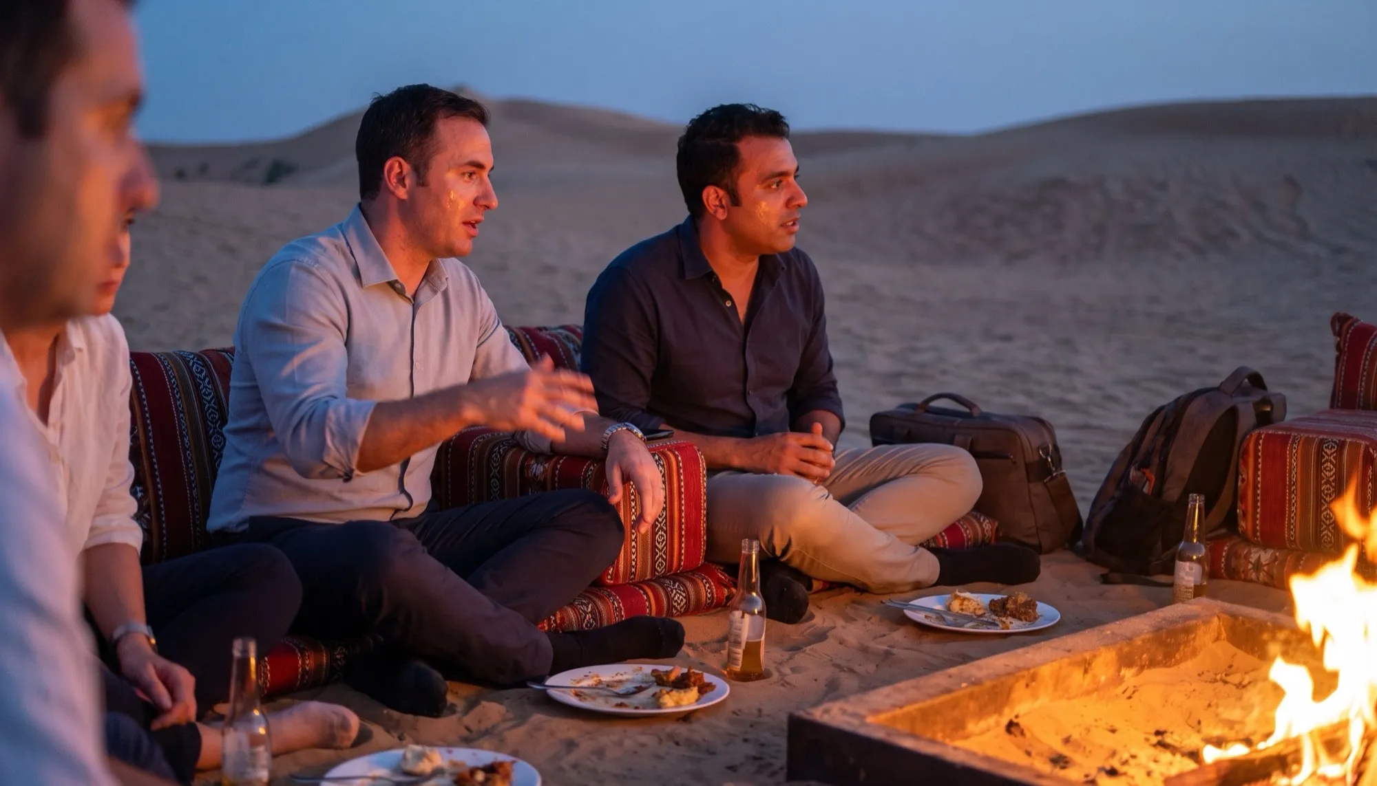 A corporate group seated on low majlis cushions around an open fire at a private desert camp at dusk, dunes behind them