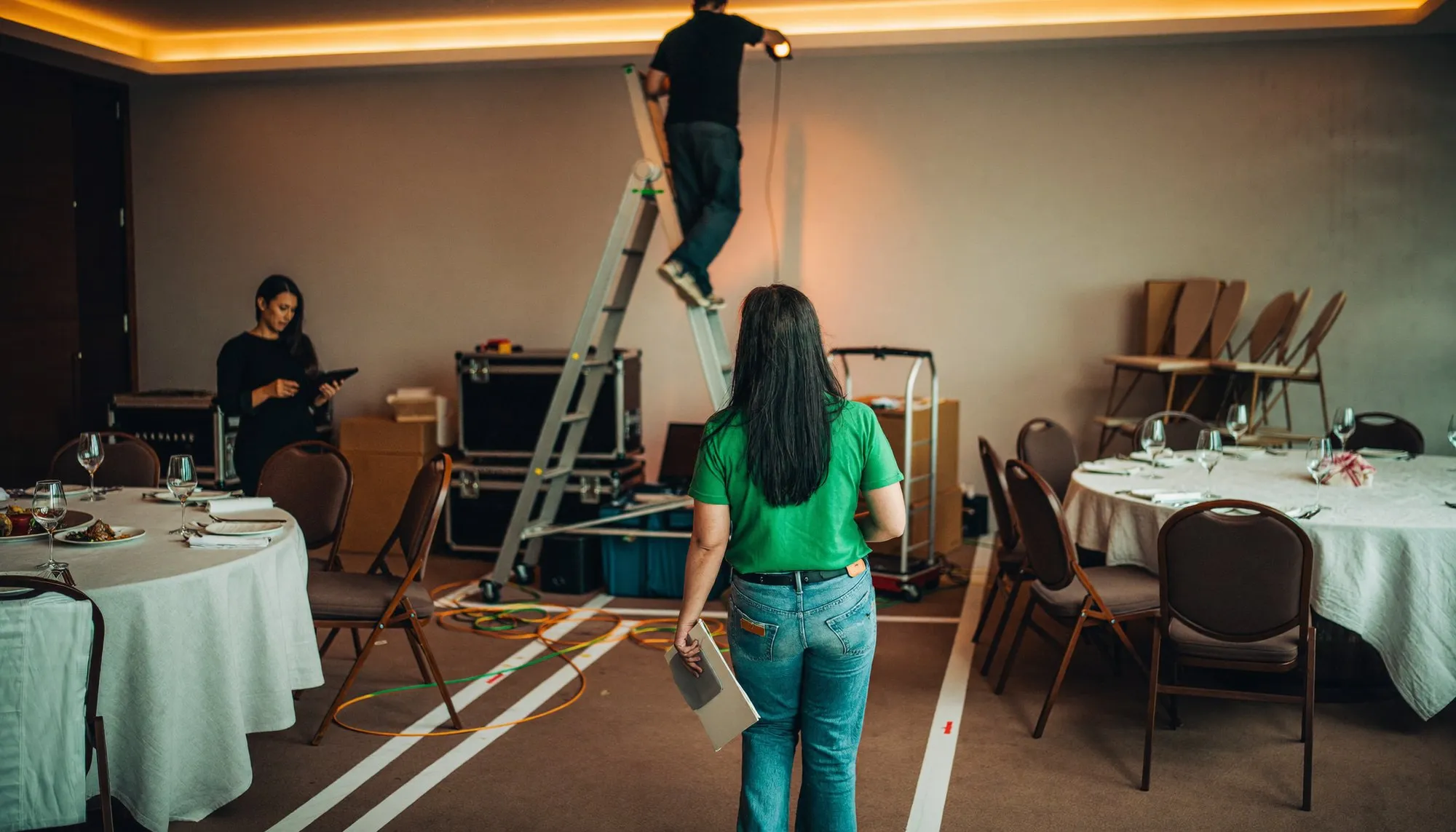A hotel function room in Dubai being set for a corporate evening, round tables half-dressed and a technician on a ladder at the back