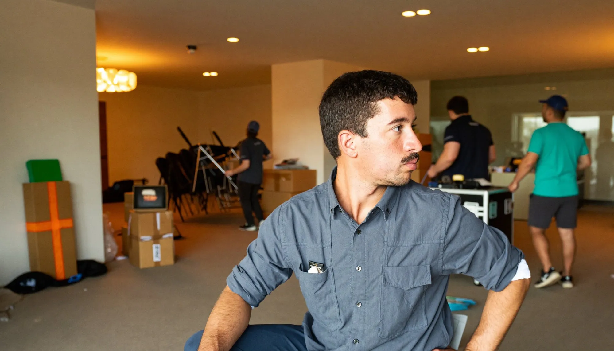 An event coordinator standing at the back of a Dubai function room during set-up, looking across it while crew move stacked chairs