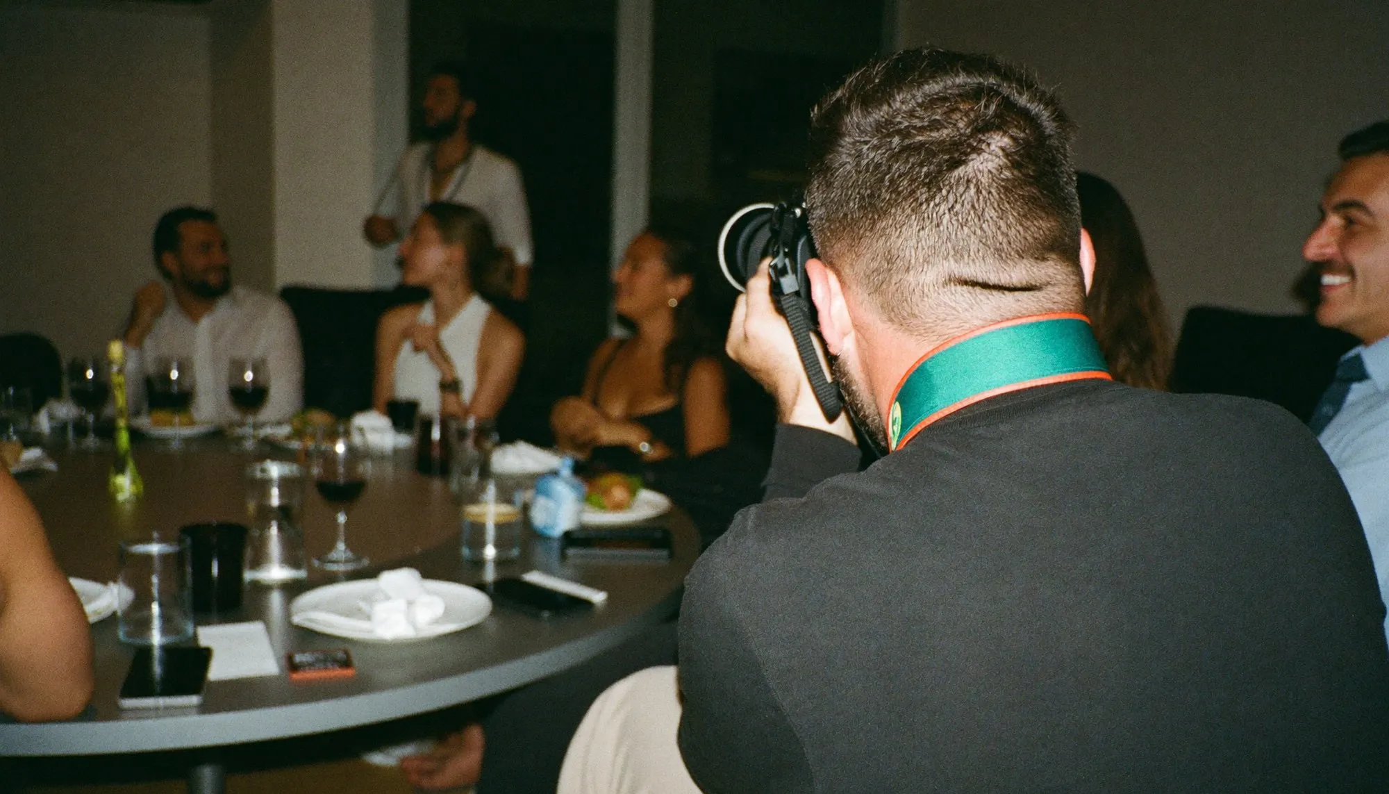 An event photographer seen from behind at a company dinner in Dubai, camera raised and pointed away, guests out of focus beyond
