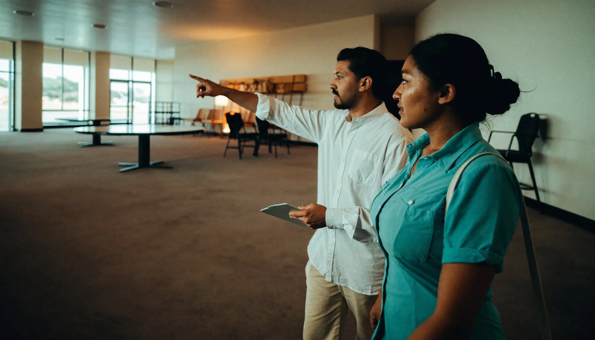 Two colleagues walking through an empty hotel function room on a site visit, one pointing across the space towards a far corner