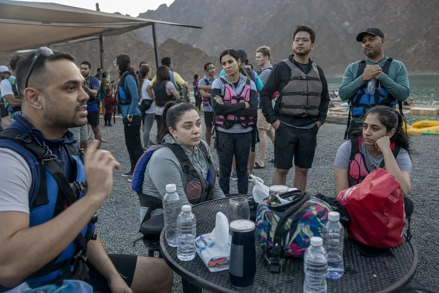 Colleagues in life jackets, leggings, shorts and caps gathered around a table of water bottles and bags at a lakeside base below rocky mountains