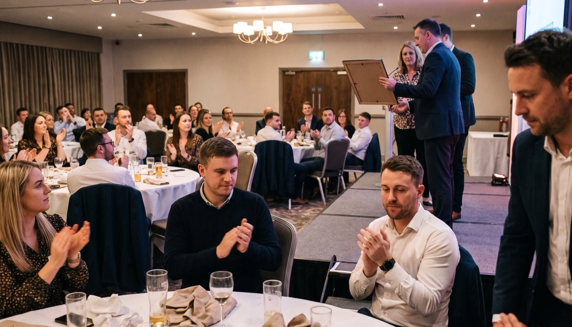 A colleague receiving a framed award on a low stage at a hotel function room while seated tables applaud