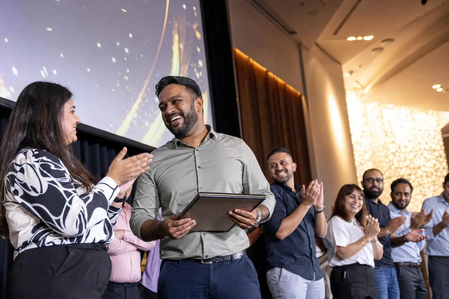A smiling employee holding a framed award on stage while a colleague and a line of team members applaud in front of a large screen