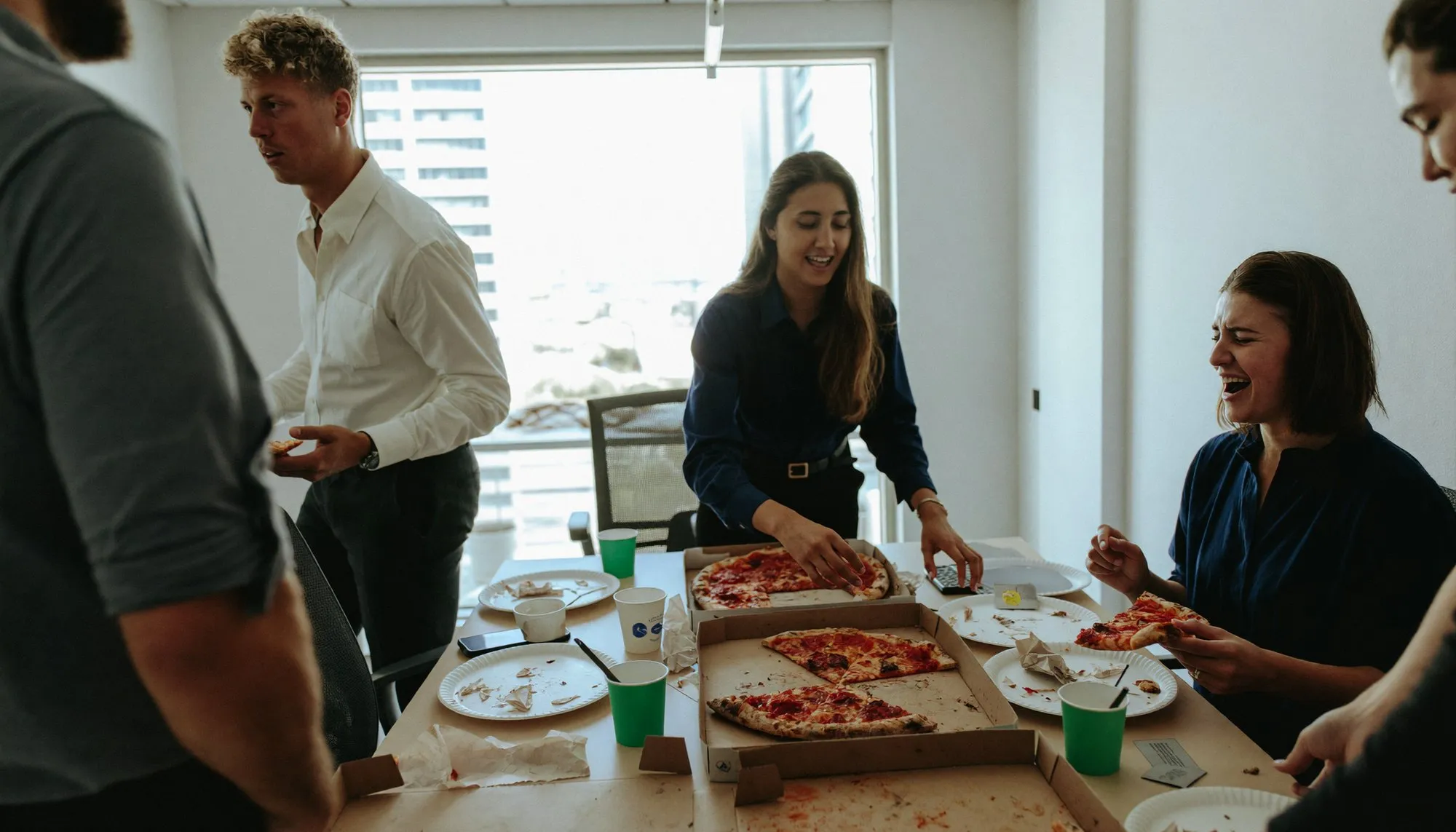 Colleagues standing around a cleared meeting table in a Dubai office with pizza boxes open, mid-conversation