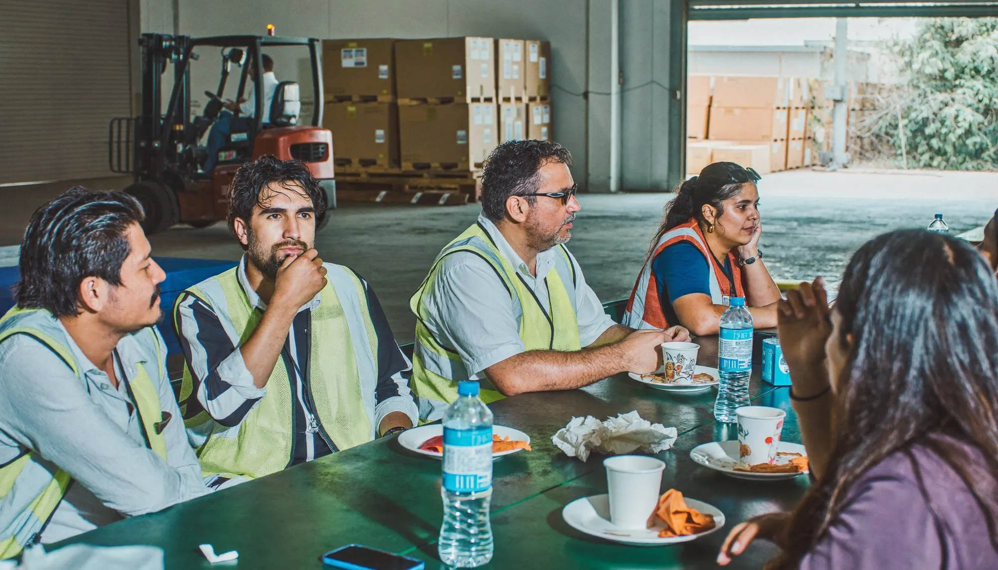 Warehouse staff in high-visibility vests sitting together at long tables during a break, plates and water bottles in front of them
