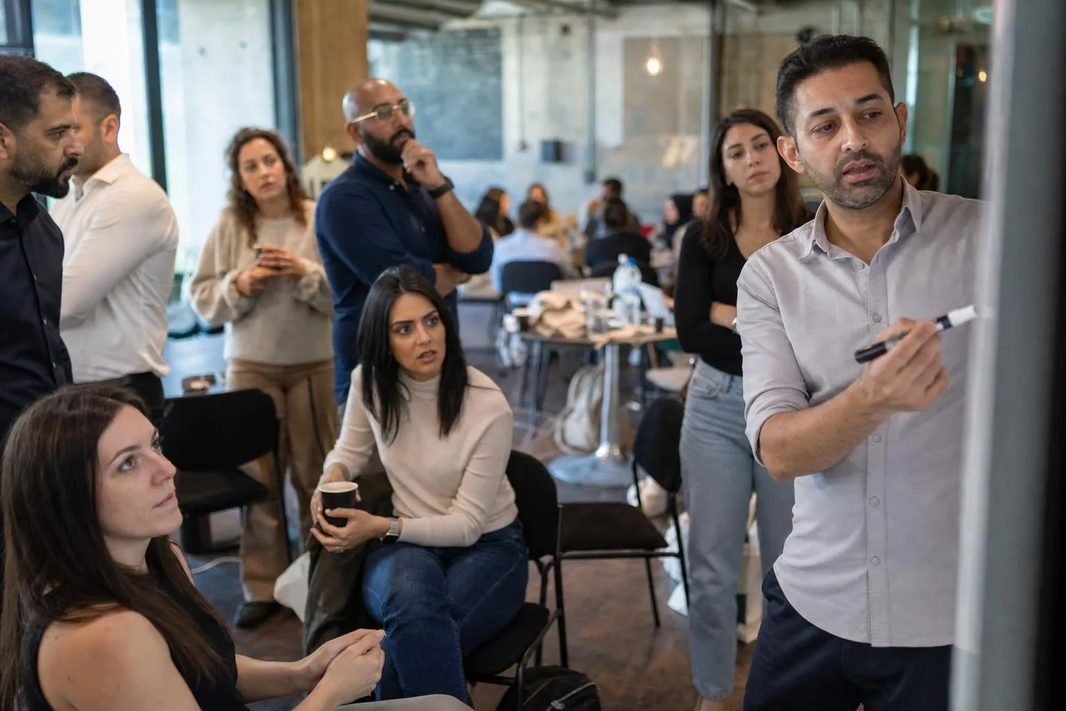 A colleague writing on a whiteboard while a mixed group of team members stands and sits around him, watching and thinking