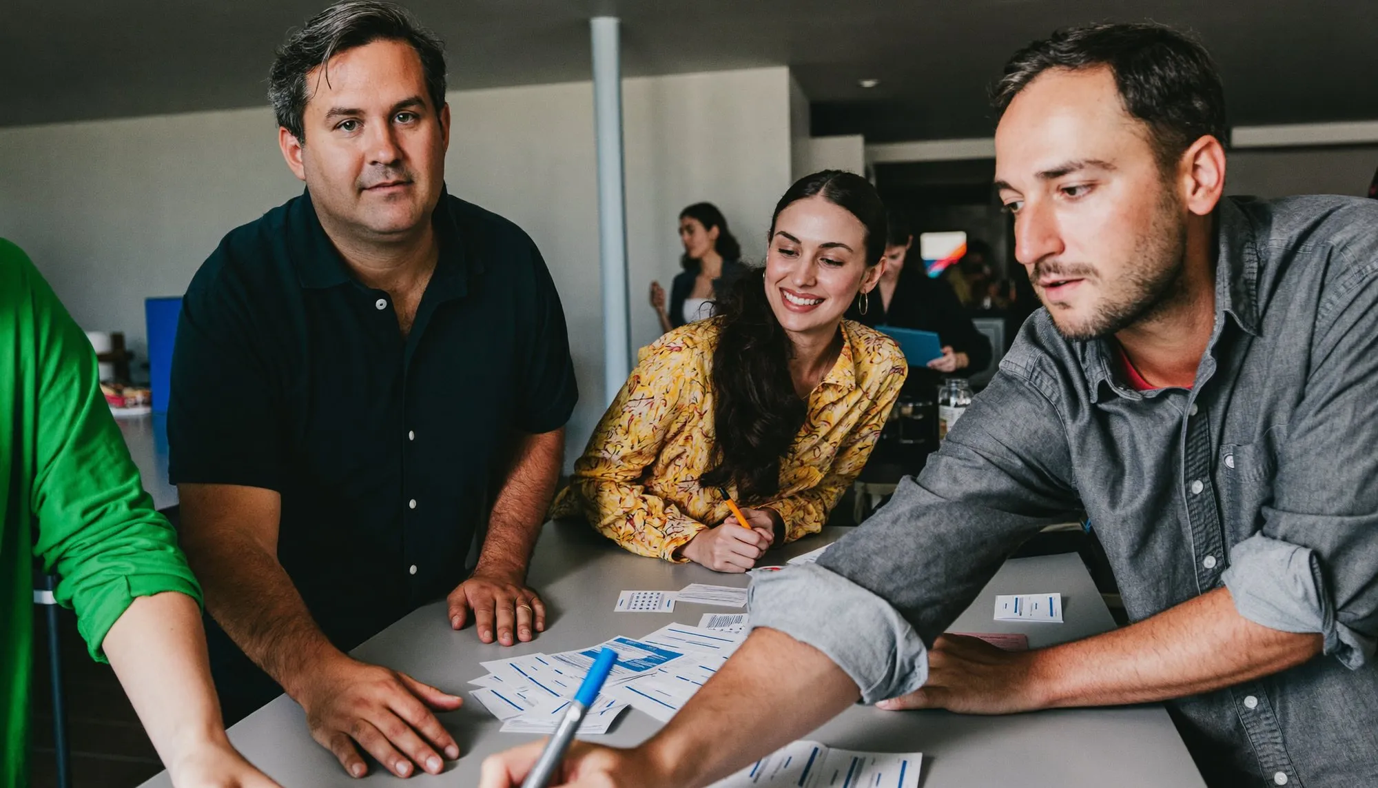 Two teams of colleagues in a bright indoor venue leaning over a table mid-task, one person reaching across with a marker