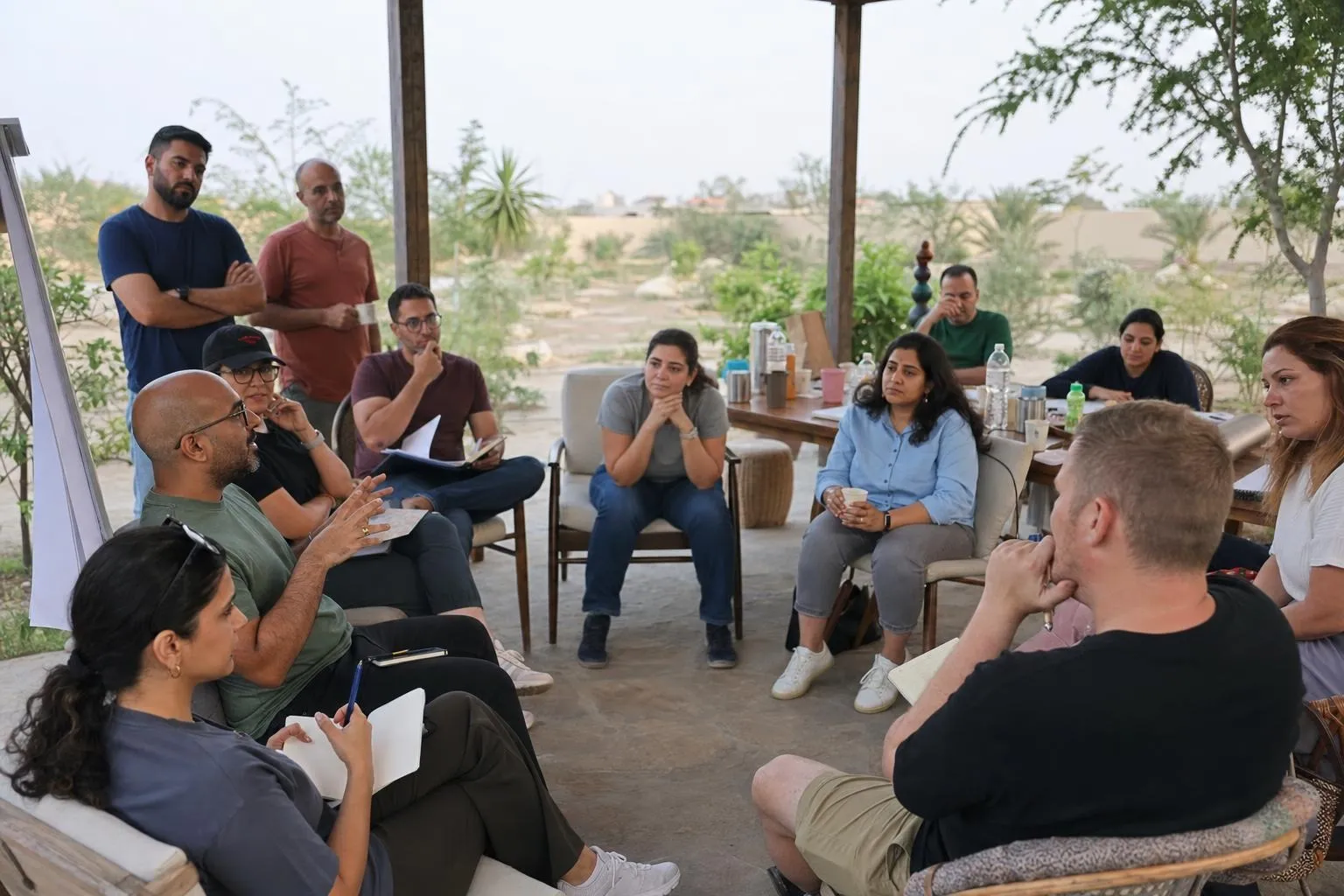 A leadership team seated in a loose circle under a shaded farmhouse terrace, notebooks open, listening to one colleague speak