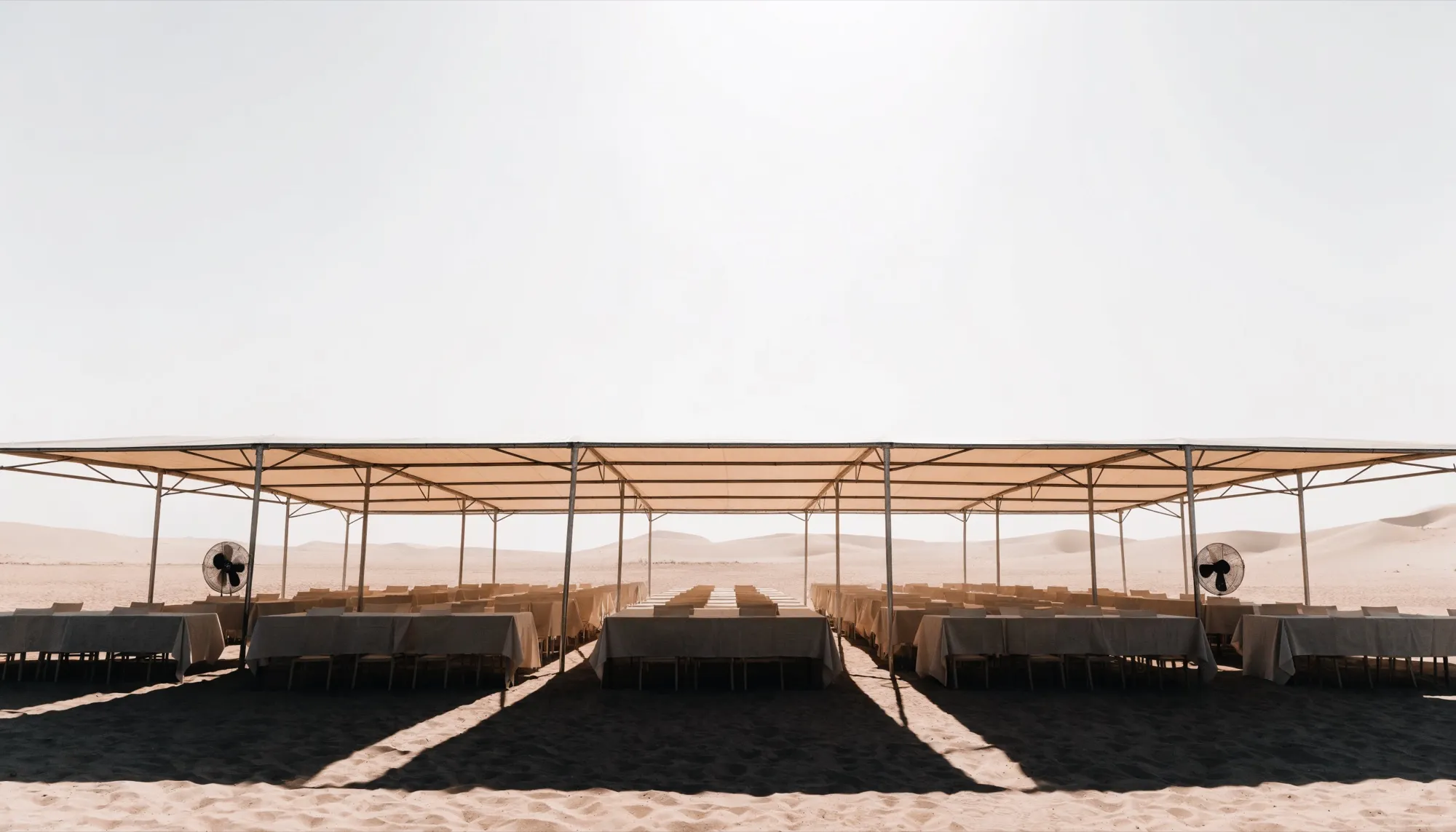 An empty shaded event marquee set on desert sand under a bleached midday sky, with heat haze over the dunes beyond